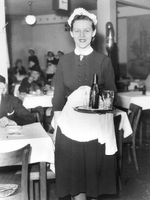 Eine Kellnerin in einem Restaurant bei der Arbeit. Auf einem Tablett serviert sie freundlich lächelnd Getränke. Aufgenommen 1953