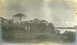 Ein Mann in Tropenkleidung sowie ein Offizier der Schutztruppe mit mehreren Afrikanern in einem Dorf an einem Fluss