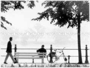 Ein Mann sitzt neben seinem Fahrrad auf einer Bank am Flussufer und ein zweiter Mann läuft vorbei