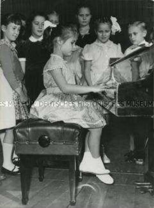 Kleines Mädchen spielt, umringt von Kindern, Klavier