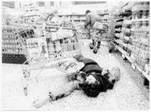 Junger Mann liegt vor einem gefüllten Einkaufswagen auf dem Supermarktboden (Altersgruppe 18-21)