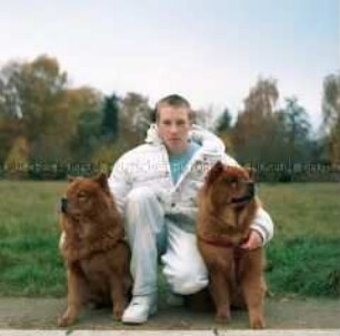 Ein Junge kniet zwischen zwei Chow-Chow-Hunden, im Hintergrund Wiese und Bäume (Altersgruppe C)