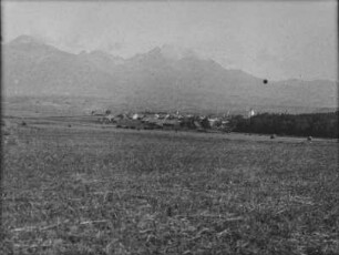 Dorf : Blick über ein abgeerntetes Feld auf ein Dorf. Im Hintergrund hohe Berge