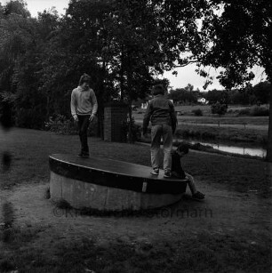 Kinderspielplatz: am Travewanderweg: drei Jungen auf Drehscheibe: Trave: am gegenüberliegenden Ufer Wiesen, Bungalow