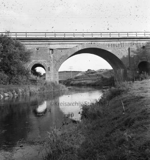 Trave: rechts und links am Ufer Böschung: hinten Brücke der Eisenbahnstrecke zwischen Hamburg und Lübeck: dahinter Wiesen