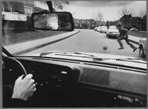 Kind mit Ball auf einer Straße vor einem Auto