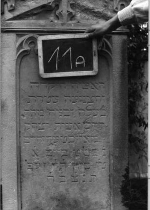 Grabstein 11 (Index: 99936899) Material: Sandstein Zustand: verwittert und beschädigt mit Textverlust Maße HxBxT: 168x56x28 cm Beschreibung: Stele, profilierter Sockel, giebelartiger Abschluß mit Verdachung mit Volutenaufsatz mit Mittelakroter, Schriftfeld eingetieft Symbolik: Maßwerk, gestelztes Rundbogenfries: Grabstein 11A