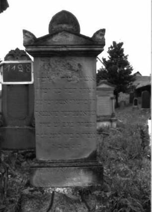 Grabstein 1198 (Index: 100104361) Material: Sandstein Zustand: stellenweise stark verwittert, teilweise noch gut erhalten Maße HxBxT: 178x47x16 cm Beschreibung: Stele auf getrepptem Sockel, Giebelaufsatz mit Giebel- und Eckakroterien, mit eingeritzten Voluten/Blattornamenten, unterster Sockel felsartig gearbeitet Symbolik: Voluten-/Blattornamente: Grabstein 1198