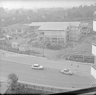 Evangelisches Gemeindehaus: Masurenweg: Rohbau: Blick von einem Hochhaus auf die Baustelle mit Bauwagen, Erdhaufen, Bretterstapel, Förderband und Baumaterial: vorn zwei Pkw auf Straße: hinten Einfamilienhäuser, Gärten und Bäume: im Hintergrund links Turm der Peter-Paul-Kirche