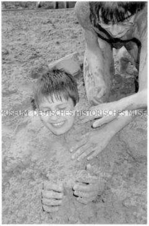 Junge wird von einem anderen Jungen im Sand eingebuddelt (Altersgruppe bis 14)