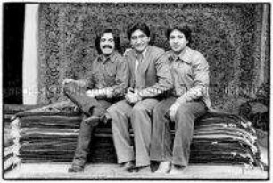 Drei Teppichhändler sitzen auf einem Stapel Teppiche (Altersgruppe 18-21)