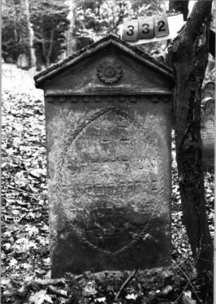 Grabstein 332 (Index: 99752334) Material: Sandstein Zustand: verwittert mit Textverlust Maße HxBxT: 128x65x14 cm Beschreibung: Stele, giebelartiger Abschluß mit Verdachung Symbolik: Blüte, Zahnfries, Lorbeerkranz um die Inschrift: Grabstein 332