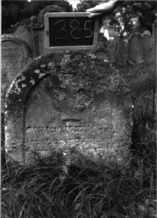 Grabstein 385 (Index: 100102852) Material: Sandstein Zustand: stark verwittert, eingesunken, weitgehender Textverlust Maße HxBxT: 77x62x10 cm Beschreibung: Stele, Rundbogenabschluss, Schriftfeld eingetieft, im Bogen Levitenkanne Symbolik: Levitenkanne: Grabstein 385