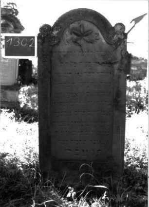 Grabstein 1302 (Index: 100104567) Material: Sandstein Zustand: leicht, oben stark verwittert Maße HxBxT: 146x64x10 cm Beschreibung: Stele mit eingezogenem Rundbogenabschluss, auf den Einzügen Kugelaufsätze, Schriftfeld eingetieft mit Konvexbogenabschluss, im Bogenfeld Blüte Symbolik: Blüte: Grabstein 1302