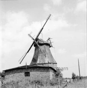 Windmühle mit beschädigten Flügeln: davor Wirtschaftsgebäude: rechts Zaun und Strommast
