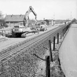 Bahnübergang bei Schrankenposten 60, Im Seybeck: Bahnstrecke zwischen Hamburg und Lübeck: Gleiserneuerung: Raupenbagger, Schaufellader, Schienenstücke, Arbeiter: hinten Schrankenwärterhaus, Schranke geöffnet, Personen auf Übergang: hinten links Wohnhaus, Garten: rechts Zaun, Straße