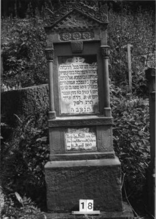 Grabstein 18 (Index: 100079843) Material: Sandstein, weiße Marmorplatten Zustand: Stele verwittert, Platten gut Maße HxBxT: 188x56x22 cm Beschreibung: ädikulaartige Stele mit Säulen, getreppter Sockel, giebelartiger Abschluß mit Eckakroteren, zwei rechteckige Schriftplatten Symbolik: Blüten, Blätter: Grabstein 18