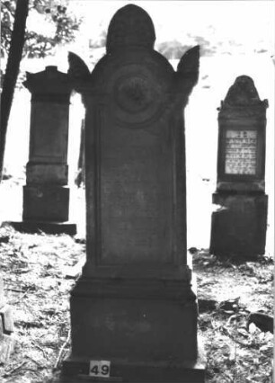 Grabstein 49 (Index: 99898779) Material: Sandstein Zustand: fast vollständig verwittert Maße HxBxT: 209x60x30 cm Beschreibung: aufgesockelte Stele, überkragender Giebelaufsatz mit Eckakroterien, unterer Sockel felsartig gearbeitet Besonderheit: vermutete Identifizierung nach Gräberverzeichnis: Grabstein 49