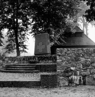 Rosenstraße / Dorfstraße: Ehrenmal mit Stufen und Feldsteinmauer: dahinter Bäume und Haus