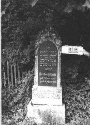 Grabstein 49 (Index: 100106560) Material: Sandstein, Schriftplatte Granit Zustand: leicht verwittert, Ornament nachgemalt Maße HxBxT: 199x53x29 cm Beschreibung: Stele mit Spitzbogenabschluß und Urnenaufsatz, Schriftplatte eingelassen, getreppter Sockel, am Bogen reiche florale Verzierung Symbolik: floral, Urne: Grabstein 49