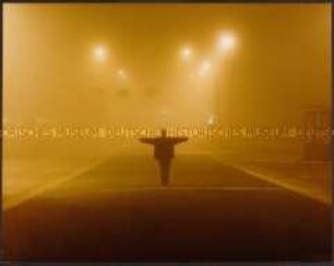 Mann steht in gelblichen, nebeligen Licht mit ausgebreiteten Armen auf einer Straße (Altersgruppe 18-21)