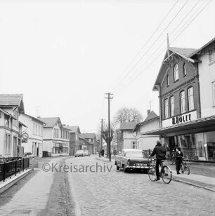 Jersbeker Straße: Blick von Hamburg Straße aus: Kopfsteinpflaster: Fahrradfahrer, parkende Autos, hinten Fußgänger: an rechter Straßenseite Strommasten, Kaufhaus Hölty: an linker Straßenseite Vorgartenzaun, Optiker, "Seegrön's Drogen Farben Fotohaus"