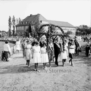 Kindervogelschießen: Umzug: vor Matthias-Claudius-Schule: Hauptkönigspaar Bärbel Rehder und Wolfgang Dreyer unter Blumenkrone, getragen von Schülerinnen: dahinter Fahne, Blumenbögen: Zuschauer, Grünanlage, Jägerzaun: im Hintergrund Häuser der Ahrensböker Straße, 27. Juni 1964