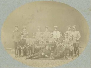 16 Fähnriche (?) in Uniform mit Mütze, teils um einen Tisch stehend, sitzend oder liegend in einem Hof, Weinflaschen und Gläser auf dem Tisch