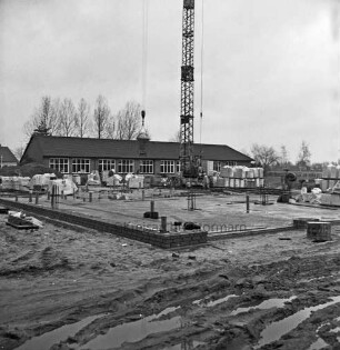 Volksschule: Erweiterungsbau: Fundament: hinten links Bauarbeiter beim Setzen der Grundmauern: rechts Stapel mit Ziegeln: in der Mitte Baukran, an Ausleger Baumaschine: vorn Furchen mit Wasserpfützen: hinten Altbau und Bäume