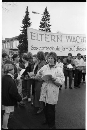 Emil-Nolde-Schule [Grundschule]: Am Schulzentrum: Elternprotest gegen Umwandlung in Gesamtschule: Brunhilde Beeken und weitere Eltern beim Verteilen von Flugblättern, Schüler, Transparent "Eltern wacht auf. Gesamtschule ja - auf diese Art nein": hinten Schulgebäude: 20. März 1990