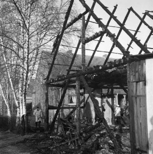 Brand: Dorfstraße: Remise mit Stall einer Nebenerwerbssiedlung: vorn rechts Brandruine, dazwischen Feuerwehrmänner: links Birken: dahinter Siedlungshäuser