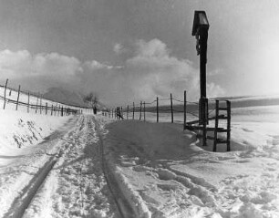 Winterbilder. Weg zwischen Koppelzäunen mit Ski- und Fußspuren. Ansicht mit Kruzifix