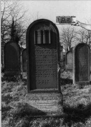Grabstein 102 (Index: 100116271) Material: Sandstein Zustand: gut Maße HxBxT: 170x54x20 cm Beschreibung: aufgesockelte Stele mit Rundbogenabschluß und eingetieftem Schriftfeld, weiteres Schriftfeld am Sockel Symbolik: aufgeschlagenes Buch mit der Aufschrift Gebetsbuch: Grabstein 102