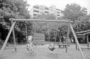 Kinderspielplatz: Kinder auf Schaukel: dahinter Bäume, Klettergerüste und Sitzgelegenheiten: im Hintergrund Hochhaus