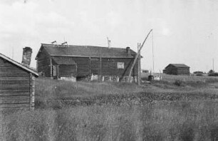 Blockhaus und Ziehbrunnen : Blockhaus in einer Siedlung. Davor ein Ziehbrunnen.
