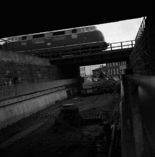 Pölitzer Weg: Bahnunterführung: Ausbau des Tunnels: Bagger beim Ausschachten unter der Unterführung: vorn Kanalisationsschacht: rechts und links Fußgängerweg mit Geländer: rechts Mann: oben Diesellok beim Überqueren des Tunnels: hinten Wohnhäuser: 16. April 1972