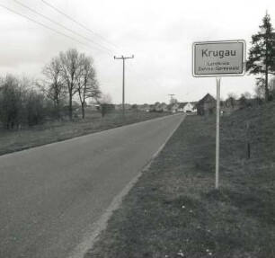 Krugau. Dorfstraße mit Ortseingangsschild