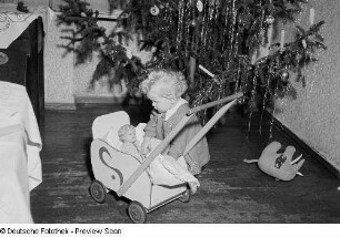 Porträt eines kleinen Mädchens mit Puppenwagen vor einem Weihnachtsbaum