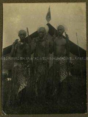 Drei alte Afrikaner in traditionellem Schmuck vor einem Zelt