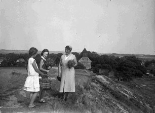 Frau mit zwei Mädchen auf einer Anhöhe, im Hintergrund eine Kirche mit kurzem Holzturm