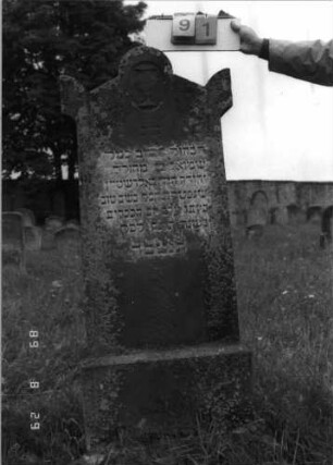 Grabstein 91 (Index: 100107298) Material: roter Sandstein Zustand: verwittert Maße HxBxT: 160x62x28 cm Beschreibung: Stele mit Giebel, Giebelfeld durch Medallion erhöht, Seitenakrotere, getreppter Sockel Symbolik: Levitenkanne: Grabstein 91