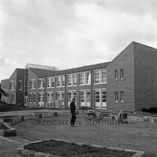 Bargteheide: Kreisgymnasium II [später Gymnasium Eckhorst]: Eckhorst: vor Fertigstellung: Herrichtung der Außenanlagen: Arbeiter beim Pflastern des Halbrunds mit Treppen und Anpflanzungen, Schubkarre: links Lüftungsrohr