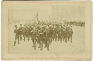 Französische Schülerkompanie in Uniform mit Gewehren und Fahne marschierend, im Hintergrund Soldaten und Zuschauer vor offentlichem Gebäude