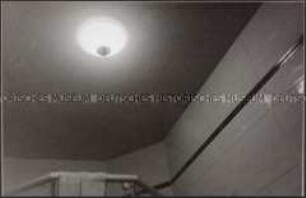 Eine runde, eingeschaltete Deckenlampe, der oberste Teil einer Duschkabine, an der Seite ist noch eine geflieste Wand mit dunkel abgesetzter Borte zu sehen