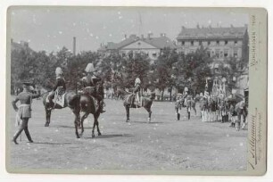 Kaiserparade auf dem Friedensplatz in Kassel im August 1899, links drei Offiziere zu Pferd, rechts Offiziere, Fähnriche mit Fahnen und Reiter, im Hintergrund Zivilbevölkerung vor Gebäuden