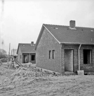 Neubausiedlung: zwei Häuser im Rohbau: hinten links Rohbau mit Dachstuhl und Gerüst: davor links Sandhaufen und Baumaterial: hinten Mann zwischen zwei Gebäuden