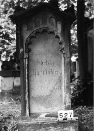 Grabstein 527 (Index: 99630028) Material: Sandstein Zustand: stark verwittert mit Textverlust Maße HxBxT: 157x68x32 cm Beschreibung: St. auf Sock. m. giebelart. Abschl. m. Verdach/Schriftfeld eingetieft m. Rundbogenabsch Symbolik: beidseitig dünne Säulen m. geschmückt. Kapitellen, Rundbogenfries, Akroterien: Grabstein 527
