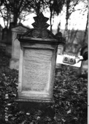 Grabstein 85 (Index: 99751842) Material: Sandstein Zustand: stark verwitt. m. fast völl. Textverlust Maße HxBxT: 158x54x24 cm Beschreibung: profil. Sock/Abdeckplatte m. Volutenaufsatz m. Palmette, rechteck. Schriftfeld einget Symbolik: Voluten, Palmette: Grabstein 85