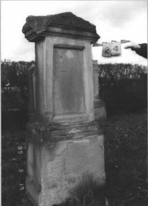 Grabstein 24 (Index: 99808545) Material: Sandstein Zustand: beschädigt, Schrifttafel fehlt, Stele und Sockel mit Zement ausgebessert Maße HxBxT: 155x59x32 cm Beschreibung: Stele, profilierter Sockel, Giebel, eingesetzte Schrifttafel (fehlend) Symbolik: Konche: Grabstein 24