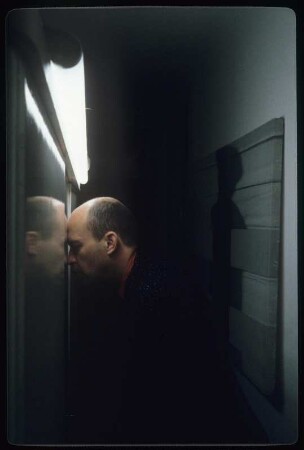 Selbstportrait Ingolf Thiel : Ingolf Thiel im schwarzen Jacket und roten Hemd in einem engen Flur, beleuchtet durch eine Neonröhre, die Stirn an einen Rahmen gelehnt; auf der anderen Wand hängt eines der Fotos aus Thiels Serie "Bandagierte Körper"
